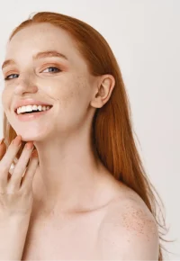 close-up-smiling-redhead-woman-with-pale-skin-freckles-touching-soft-perfect-face-using-skincare-cream-standing-white-wall1