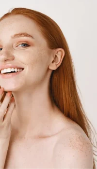 close-up-smiling-redhead-woman-with-pale-skin-freckles-touching-soft-perfect-face-using-skincare-cream-standing-white-wall1