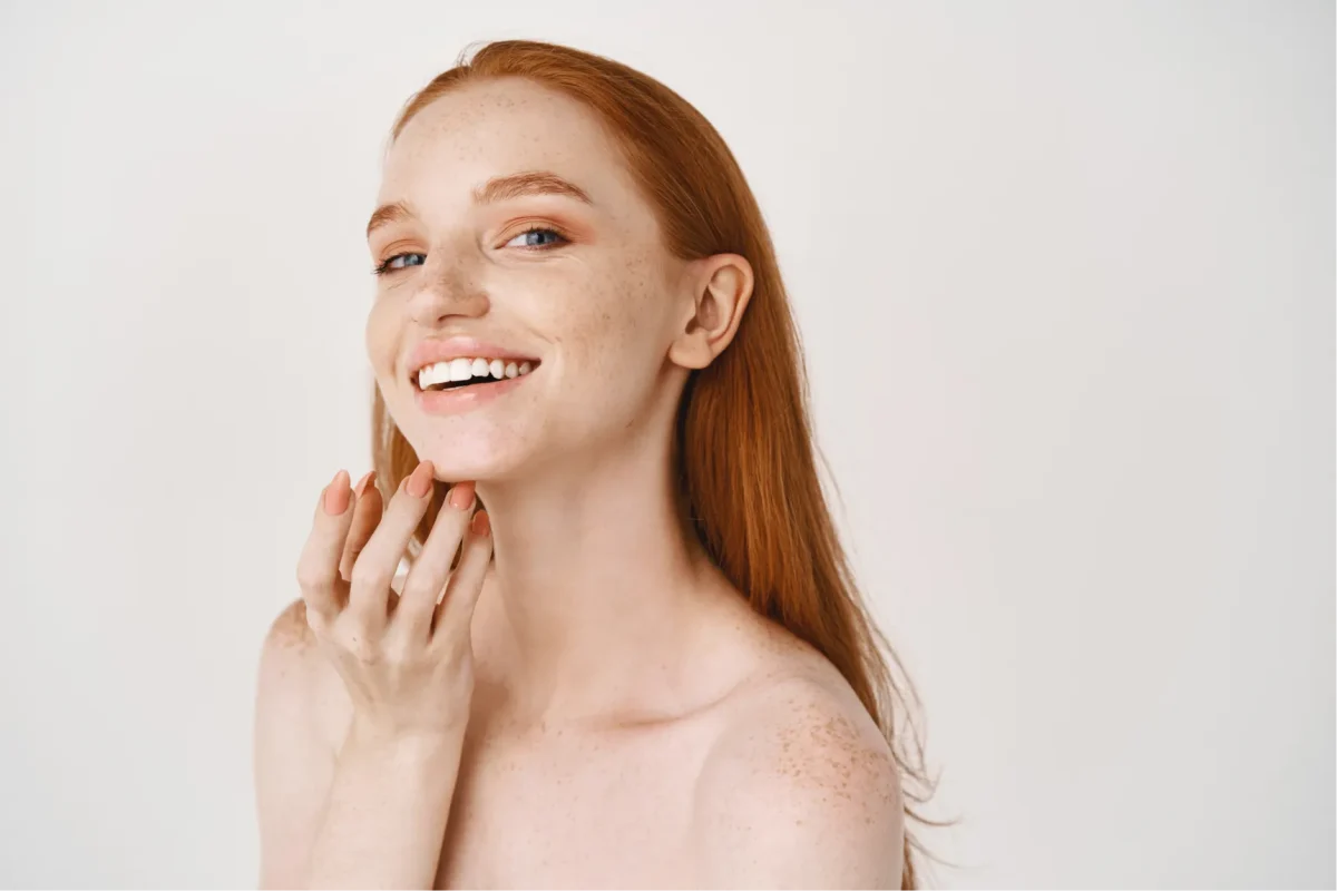 close-up-smiling-redhead-woman-with-pale-skin-freckles-touching-soft-perfect-face-using-skincare-cream-standing-white-wall1